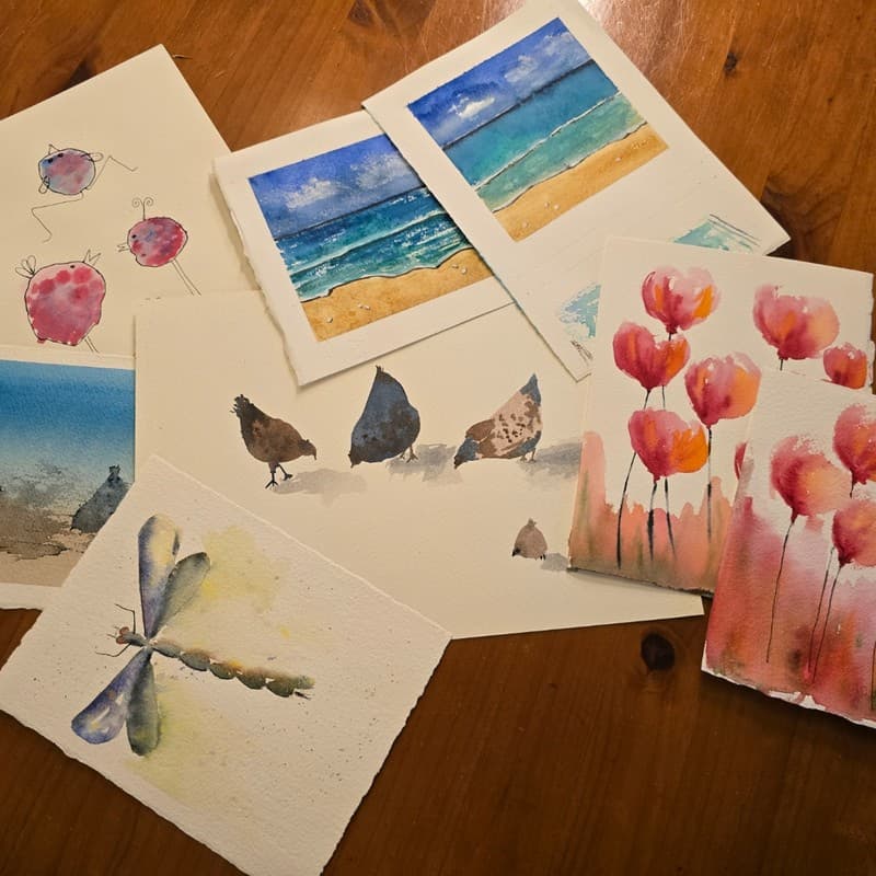 Watercolor paintings of a dragonfly, flowers, birds, and beach scenes on a wooden table.